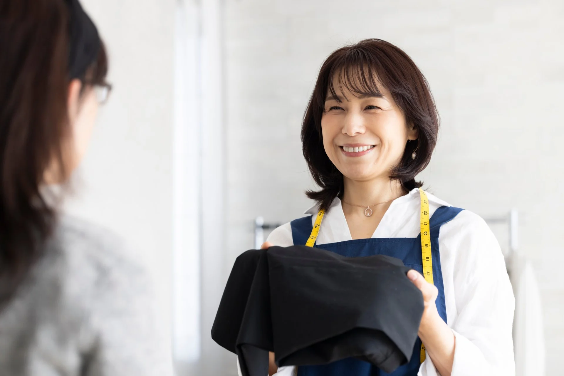 採寸テープを首にかけ、黒い衣類を手に持って笑顔で接客するクリーニング店のスタッフ